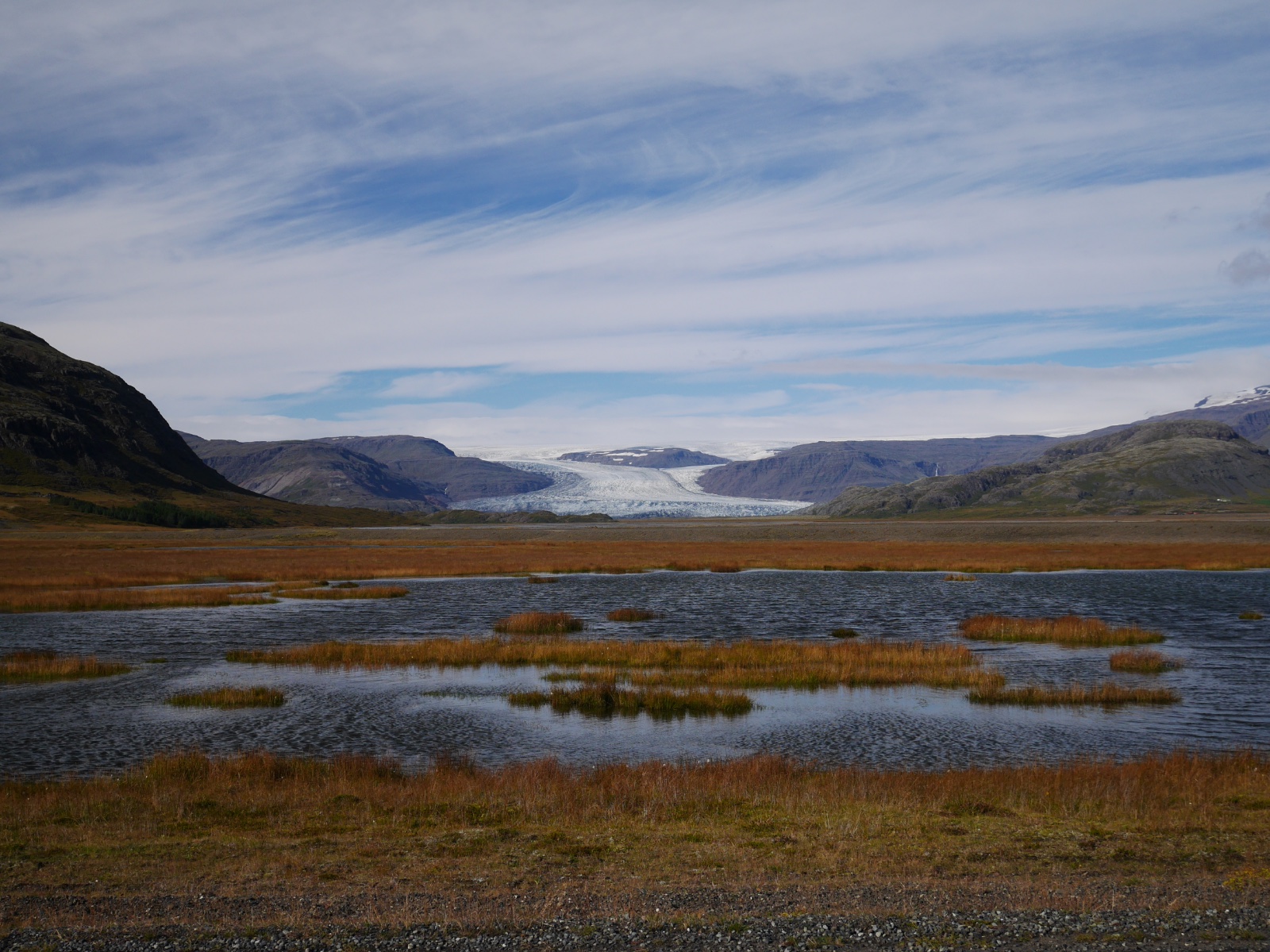 A glacier front seen across a wide outwash plain of shallow water and autumn grass.