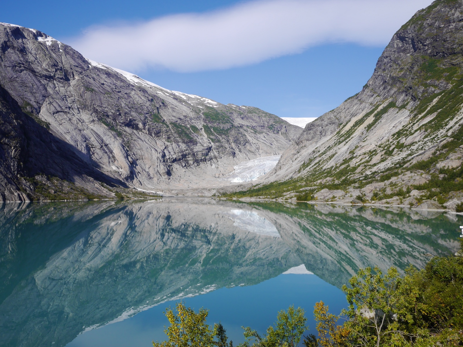 A glacier tongue descending between grey cliffs, mirrored in a still green lake.