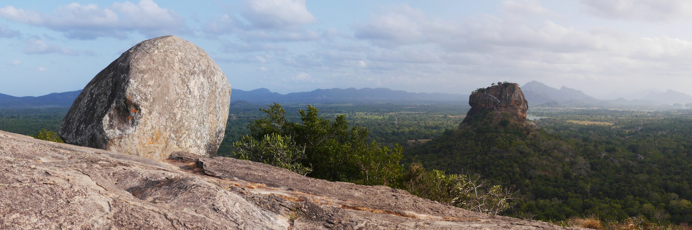 Sigiriya rock rising from forest, seen across a bare rock summit with a boulder in the foreground.